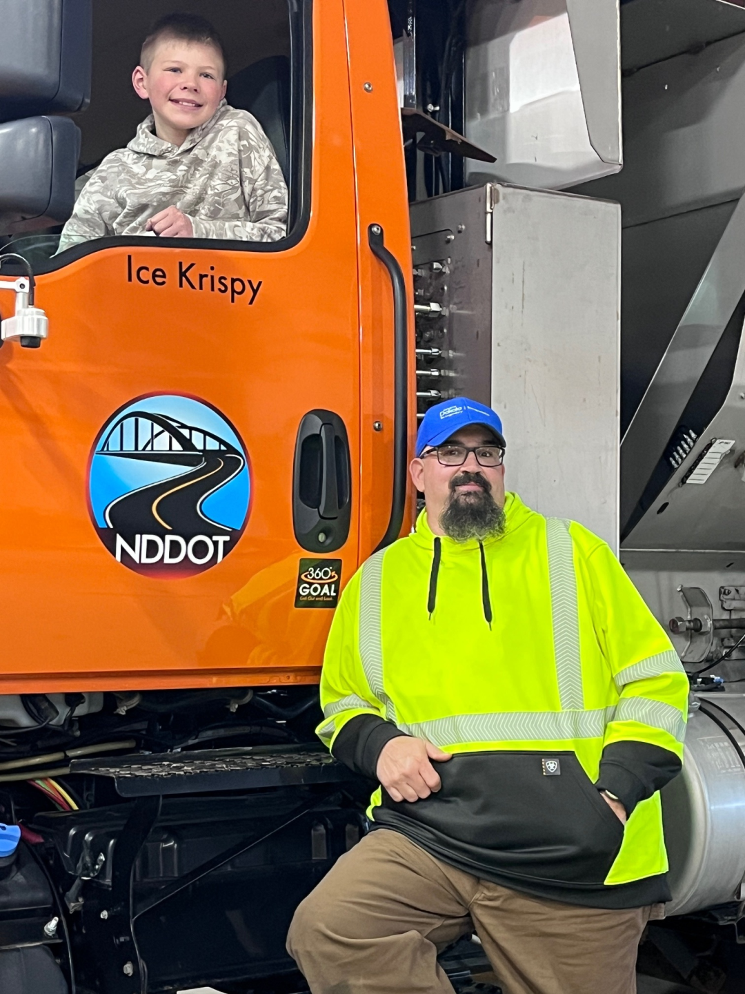 A boy looks out of a snowplow window. Ice Krispy is written on the door. A man stands beside the plow.