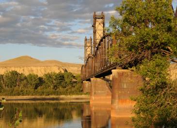 A view of a river with a railway bridge overhead at dusk.