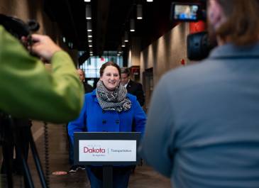 Women in blue coat and scarf stands at a podium with a NDDOT logo talking to reporters.