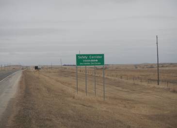 A portion of the US 85 roadway on the left with a 65 MPH speed sign and a Vision Zero road sign. Sky is gray with dry grass. 