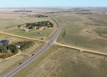 Aerial view of US 85 curving to the left through a scenic rural landscape, with nearby farmsteads, open fields, and scattered trees. The image highlights the highway’s surroundings as part of the US 85 expansion project.