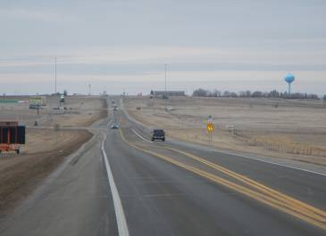View entering US 85 to Belfield, ND, showing a 55 mph speed limit sign in the foreground, a vehicle on the highway, and a gas station with a water tower visible in the distance, surrounded by dry grass.