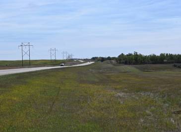 View of the US 85 Expansion project from the Junction of ND 200 North to RP 120.3, showing a rural highway bordered by green grass and powerlines, with vehicles visible in the distance under a clear blue sky.