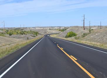 Wide-angle view of a freshly paved two-lane highway, part of the US 85 Expansion, surrounded by rolling hills, sparse vegetation.