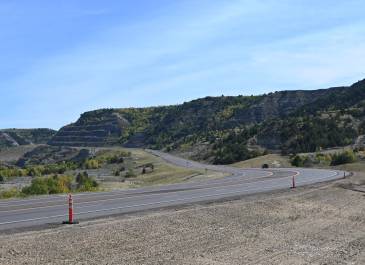 Scenic view of a winding, newly paved highway cutting through rugged hills and valleys with sparse vegetation, part of the US 85 Expansion project. Orange construction cones line the roadside under a clear blue sky.