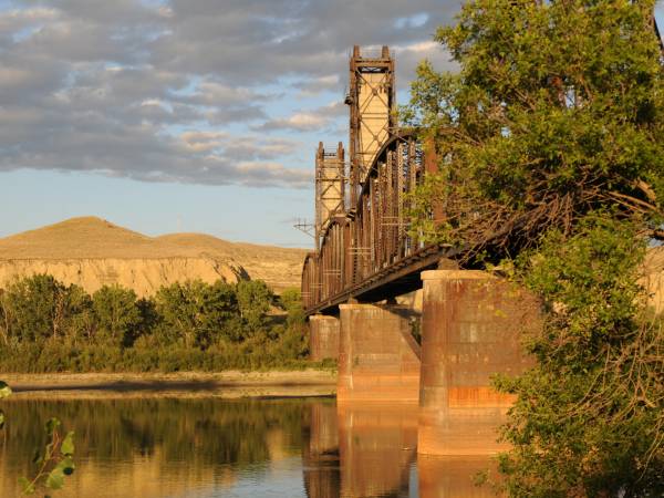 A view of a river with a railway bridge overhead at dusk.
