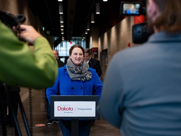 Women in blue coat and scarf stands at a podium with a NDDOT logo talking to reporters.