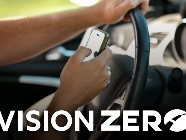 Hands holding a cell phone and texting while driving with a Vision Zero logo at the bottom.