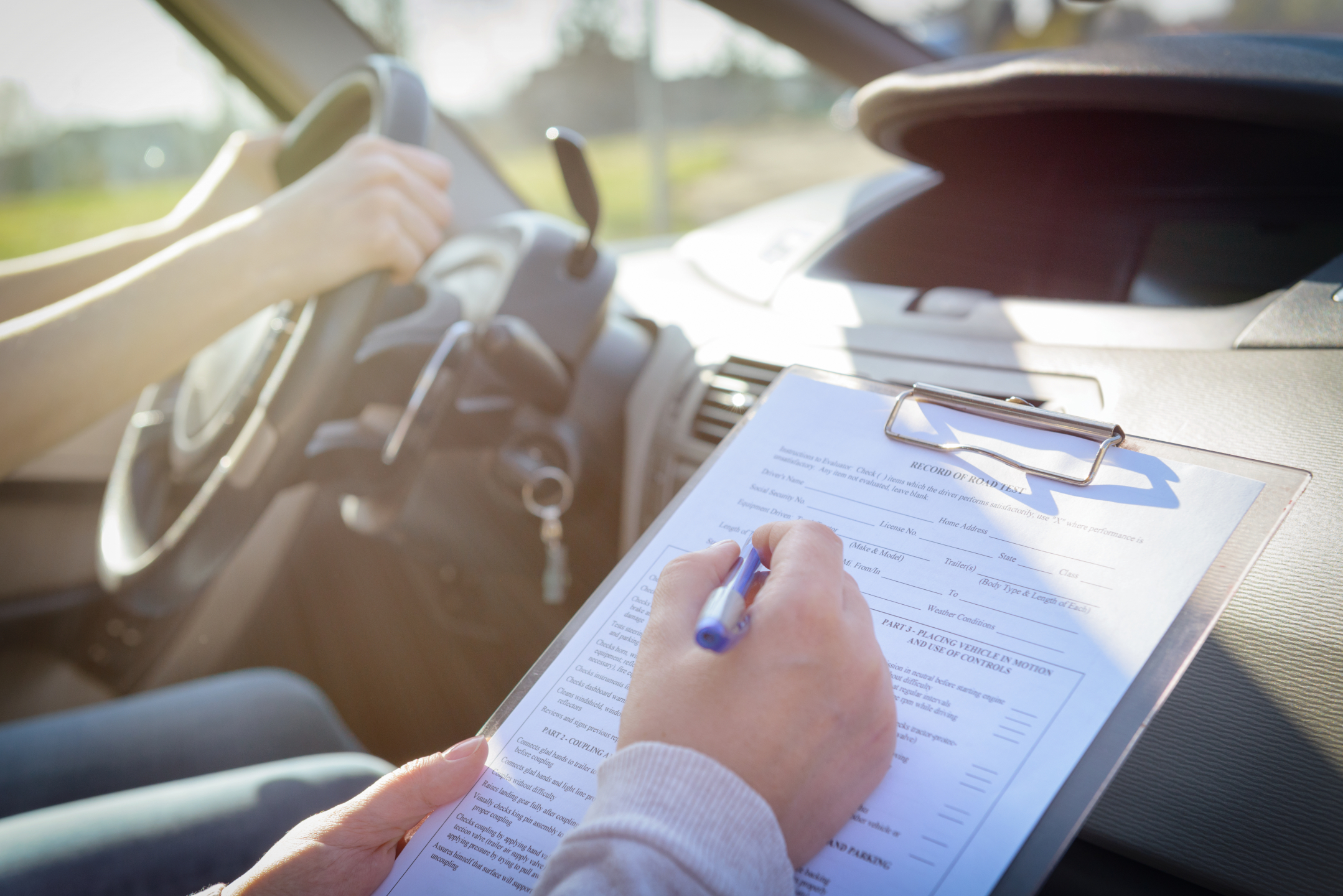 Person behind the wheel of a car while instructor in passenger seat takes notes on a clipboard