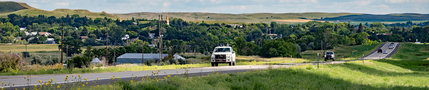 Car driving down roadway with rural residential and hills in background.