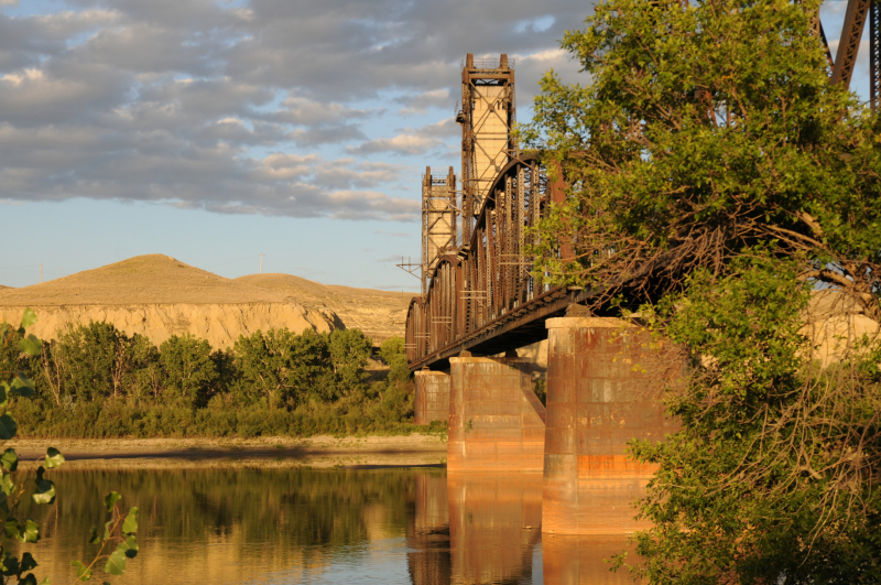 A view of a river with a railway bridge overhead at dusk.